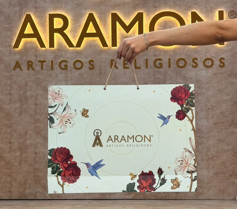 Sacola de papel personalizada com estampa floral e alça de cordão, produzida pela JK Artes Gráficas para a loja Aramon Artigos Religiosos